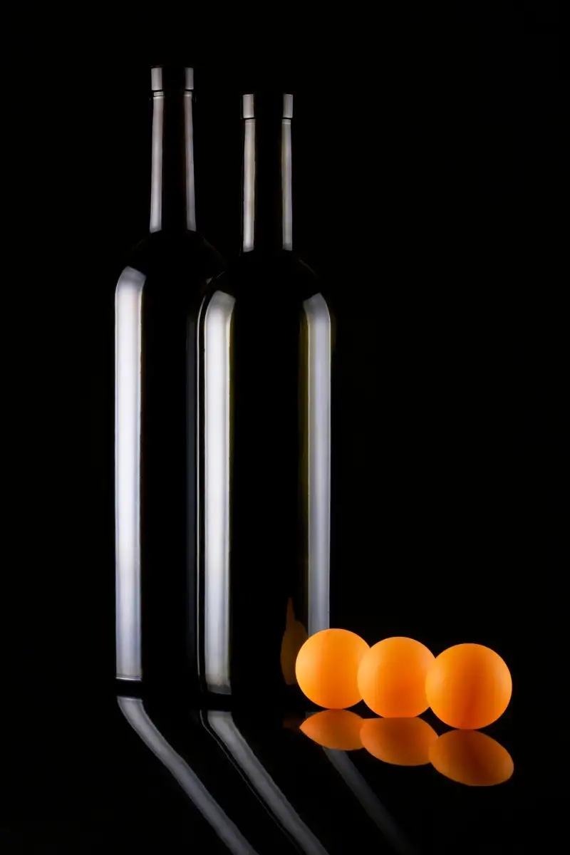 Two dark glass bottles with edge light against black, and three orange balls with their reflection