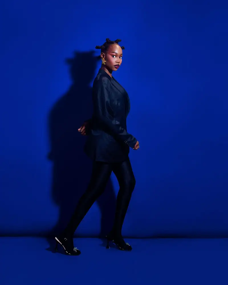 A model in a dark blazer and heels walks across a saturated blue studio backdrop, her shadow on the wall