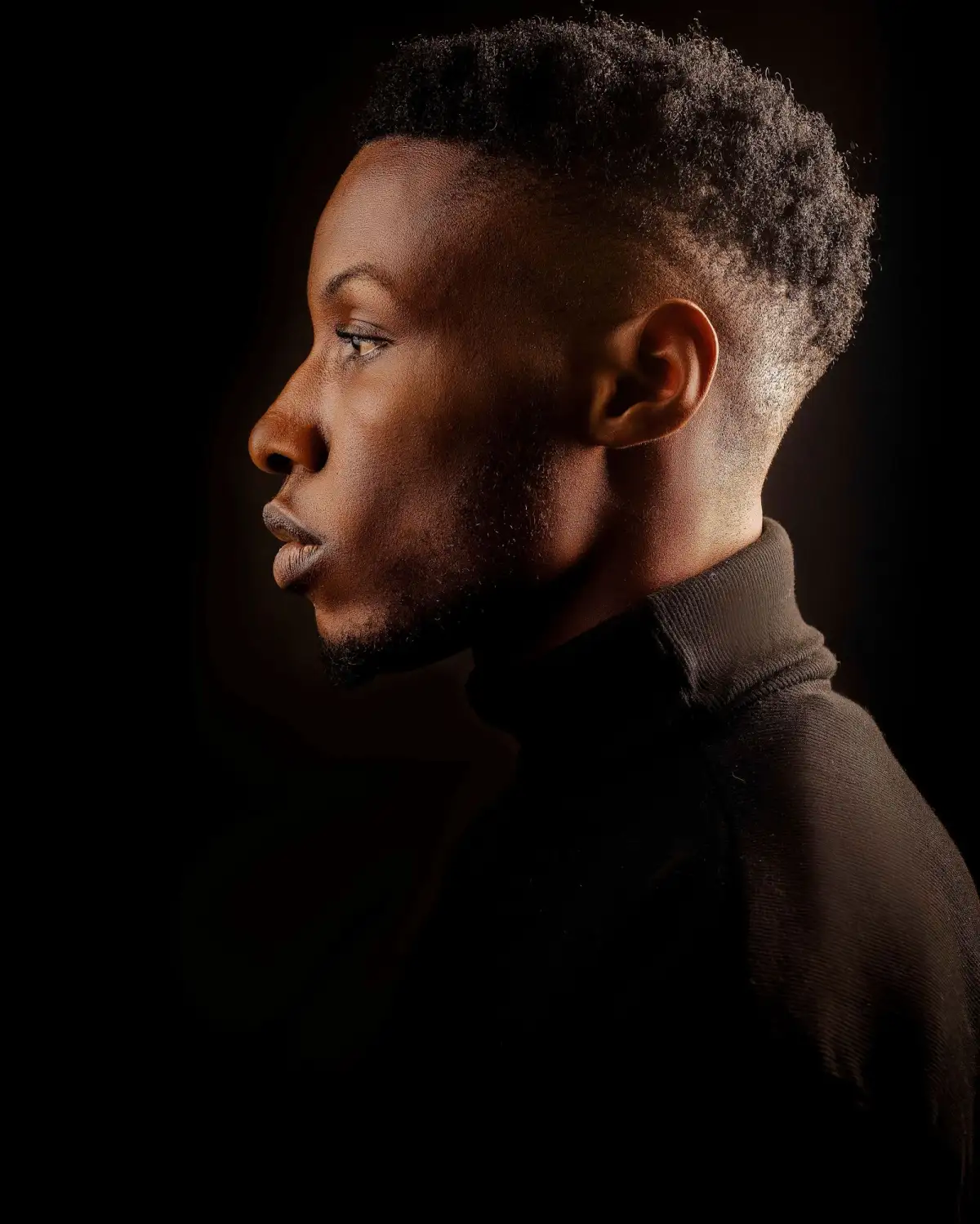 Profile portrait of a young man in a dark turtleneck, lit from the side against a black background