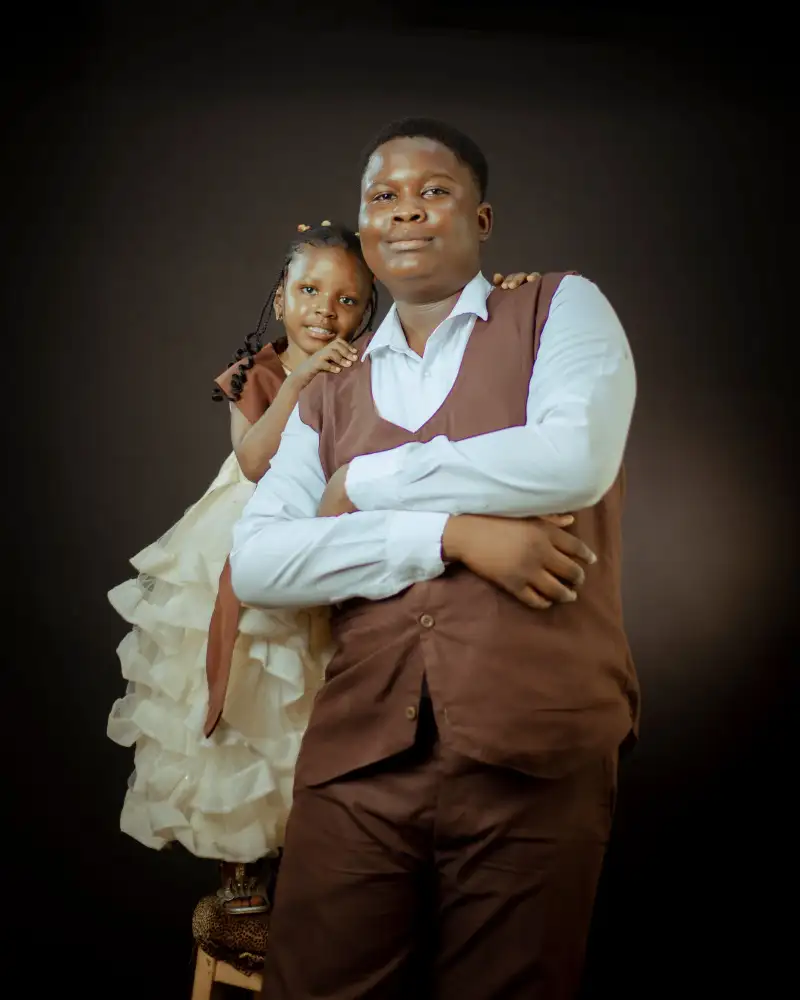 A young man in a brown waistcoat with a girl in a cream frilled dress leaning on his shoulder, against a dark studio backdrop