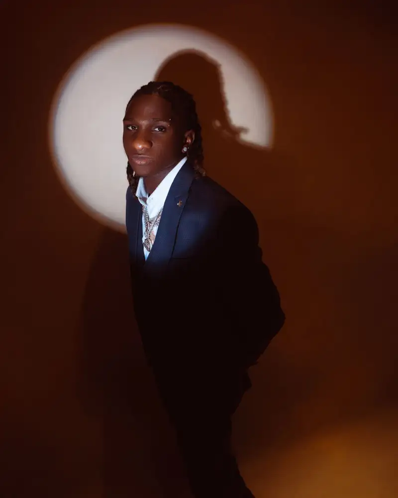A young man in a navy jacket stands in a pool of light against a warm amber wall