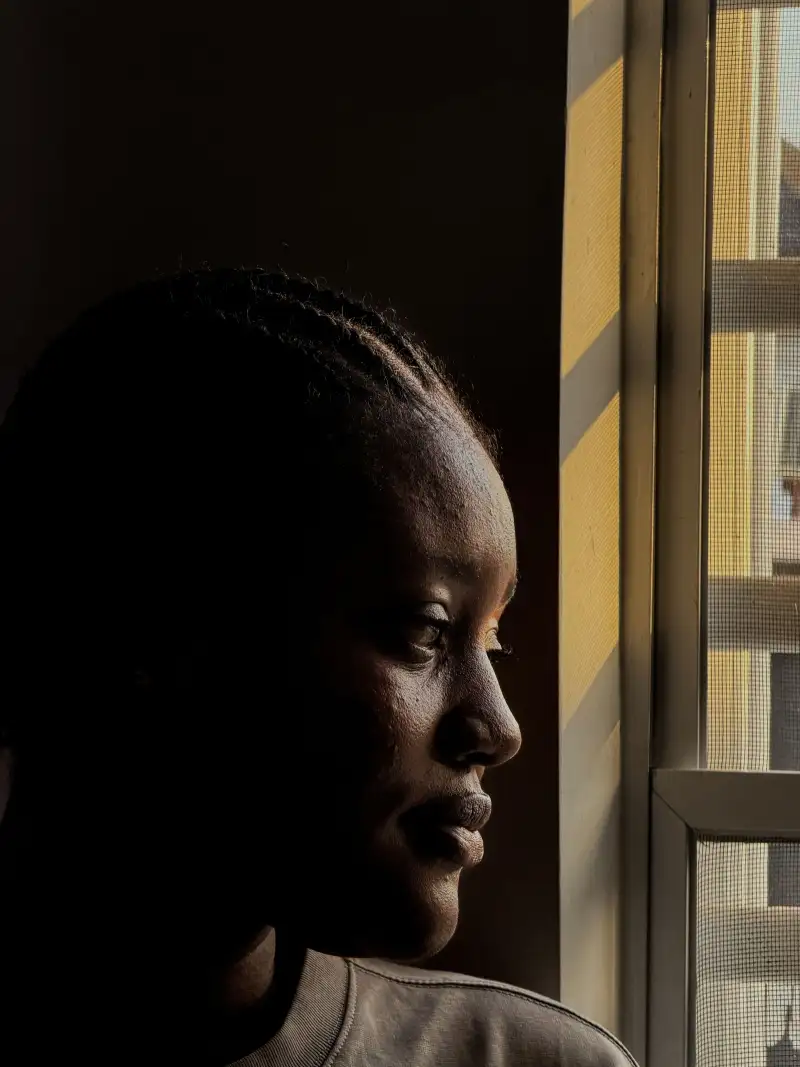 A woman with braided hair in profile, lit from a window at her side, the rest of the frame in shadow