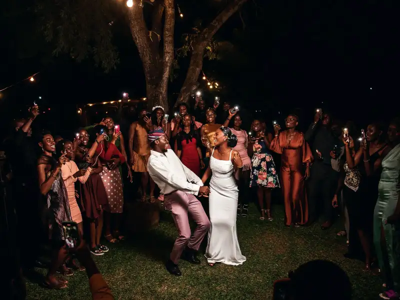 A bride and groom dance on grass at night while guests around them hold up glowing phones