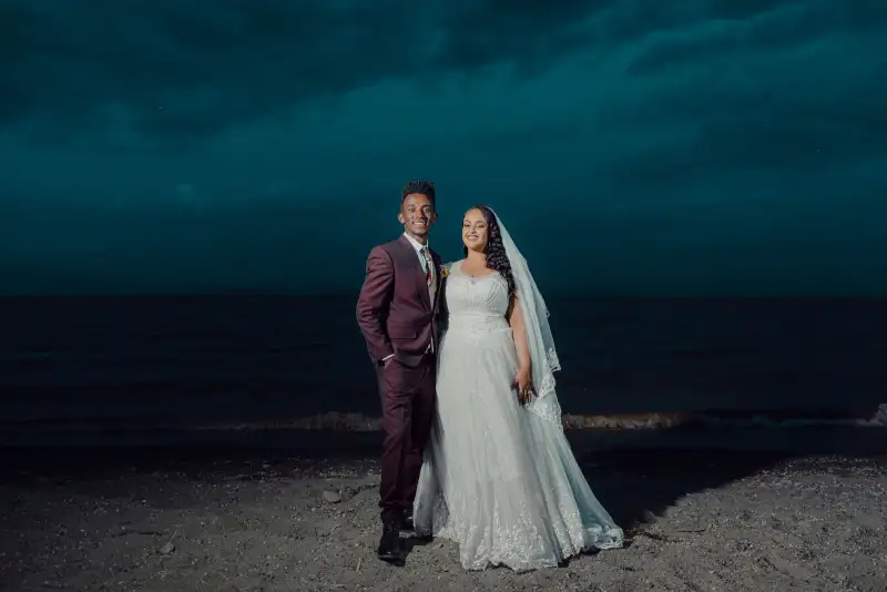 A couple in wedding clothes stand on a dark beach under a teal evening sky