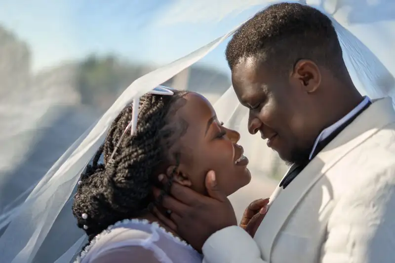 A bride and groom touch foreheads under a sheer veil in bright daylight