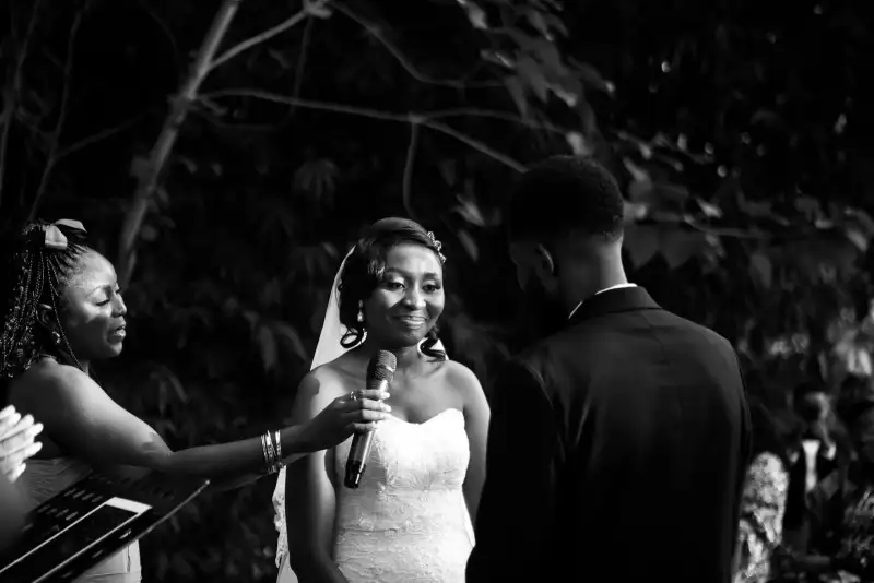 Black-and-white photograph of a bride smiling as a woman holds a microphone to her at an outdoor ceremony