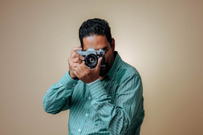 A photographer holding a camera against a warm beige backdrop