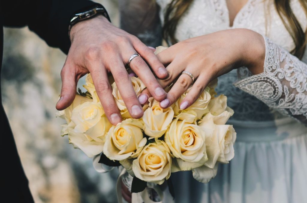 A couple's hands with wedding rings resting on a bouquet of yellow roses