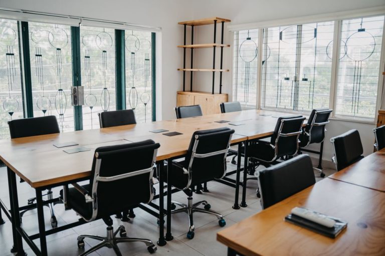 A bright meeting room with a long table, black chairs and tall windows