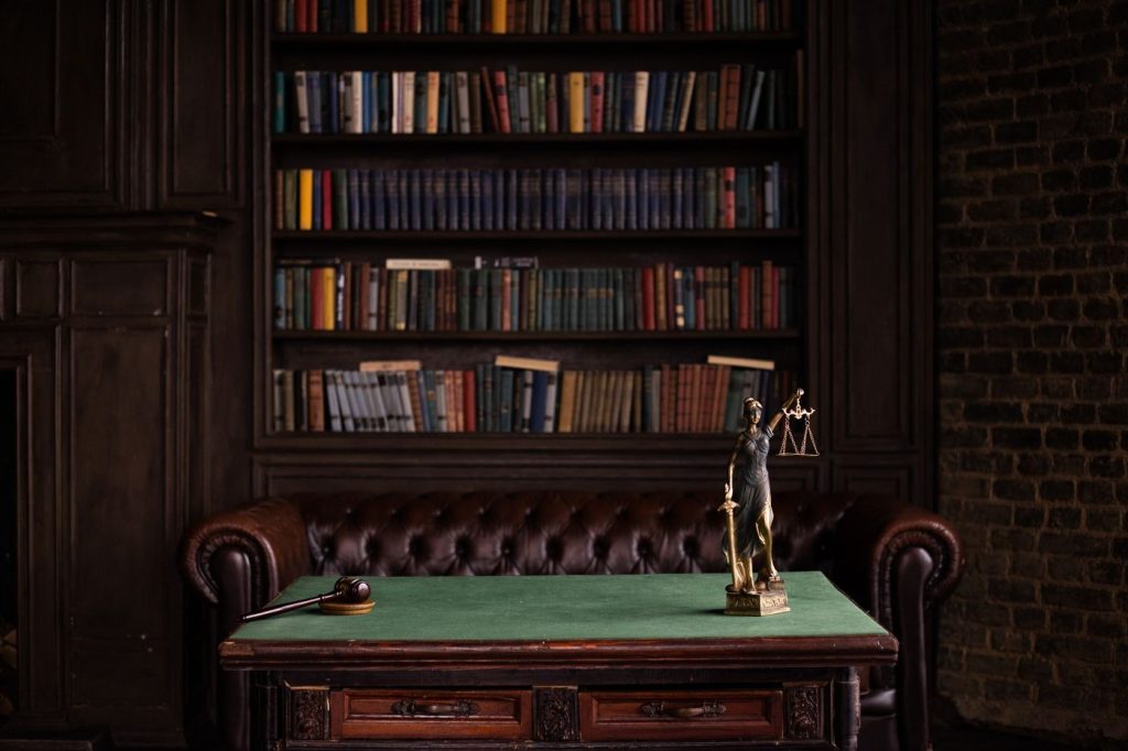 A dark wood law office with a green-topped desk, a gavel, a figure of justice and shelves of books