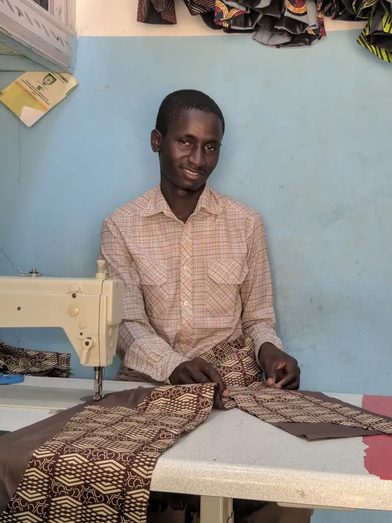 A smiling tailor in a checked shirt sitting at a sewing machine with printed cloth on the table