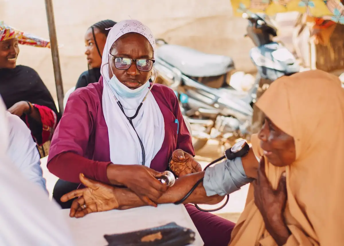 A health worker in a maroon uniform and white headscarf checks the blood pressure of an older woman at an outdoor clinic while others look on