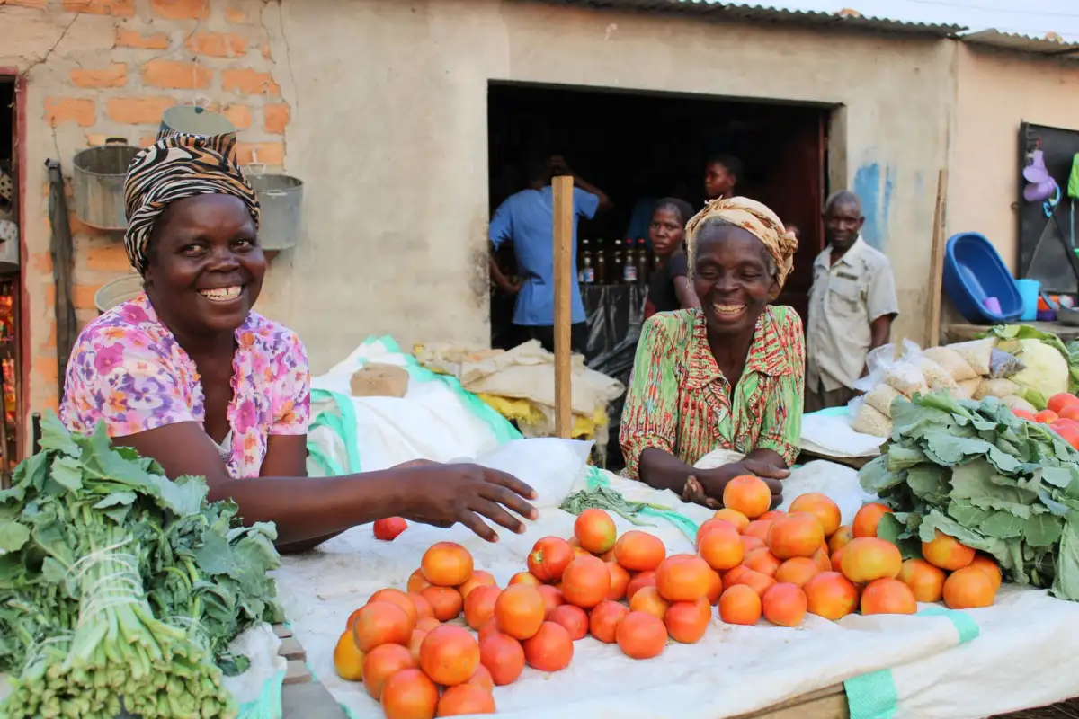 Two smiling women stand behind a market stall piled with tomatoes and leafy greens
