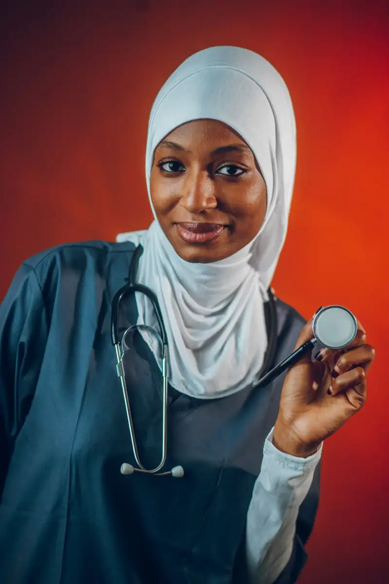 A smiling health worker in a dark uniform and light headscarf holds a stethoscope against a red background