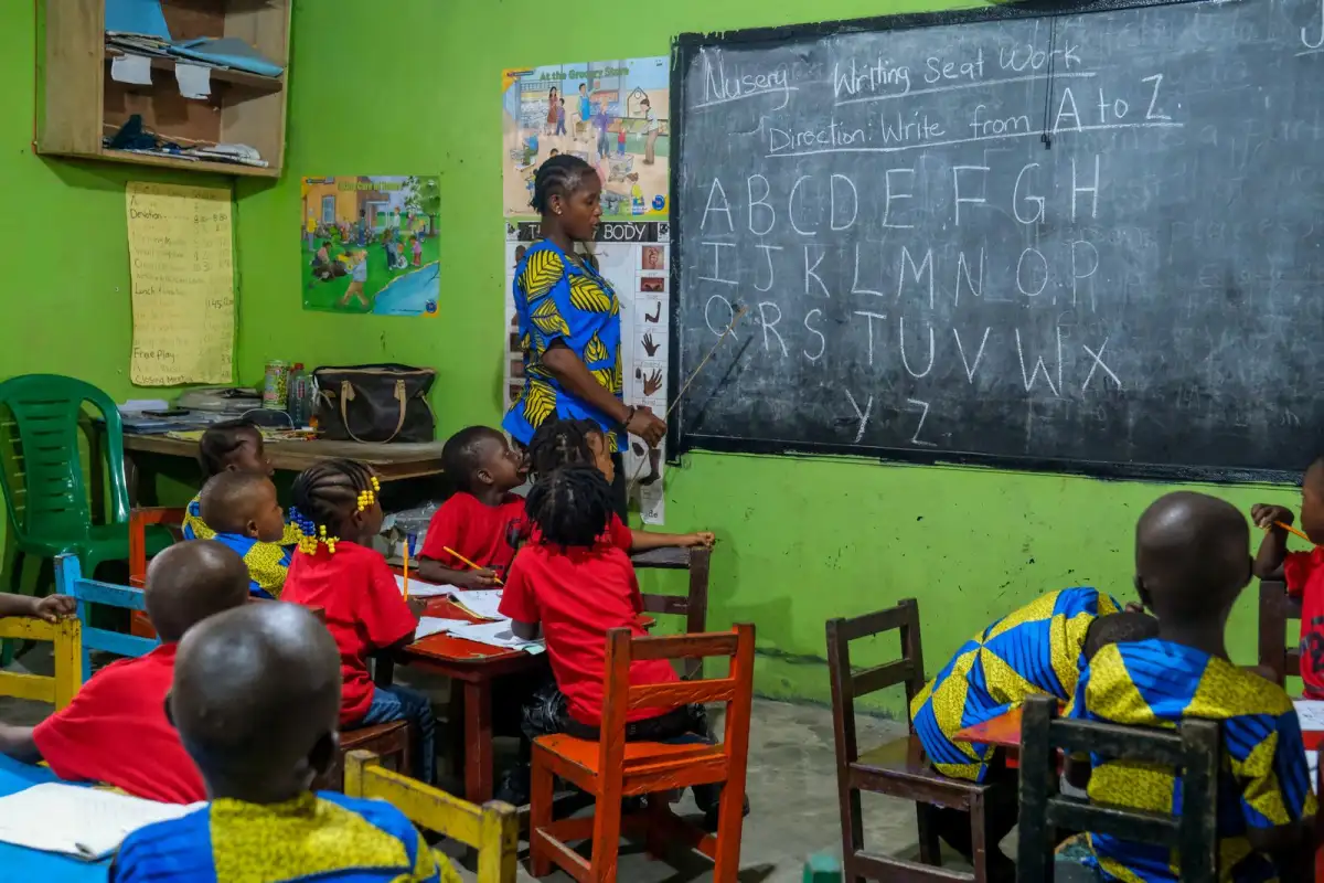 Children in red and blue sit at wooden desks in a bright green classroom with a chalkboard showing the alphabet and a teacher standing beside it