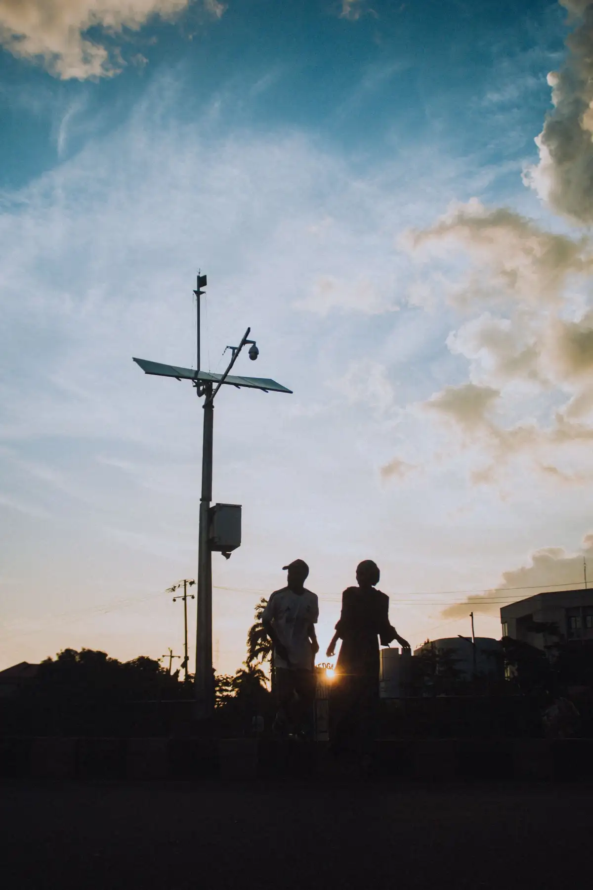 Two people walk along a road at sunset beneath a pole with a solar panel and street light, silhouetted against a cloudy sky
