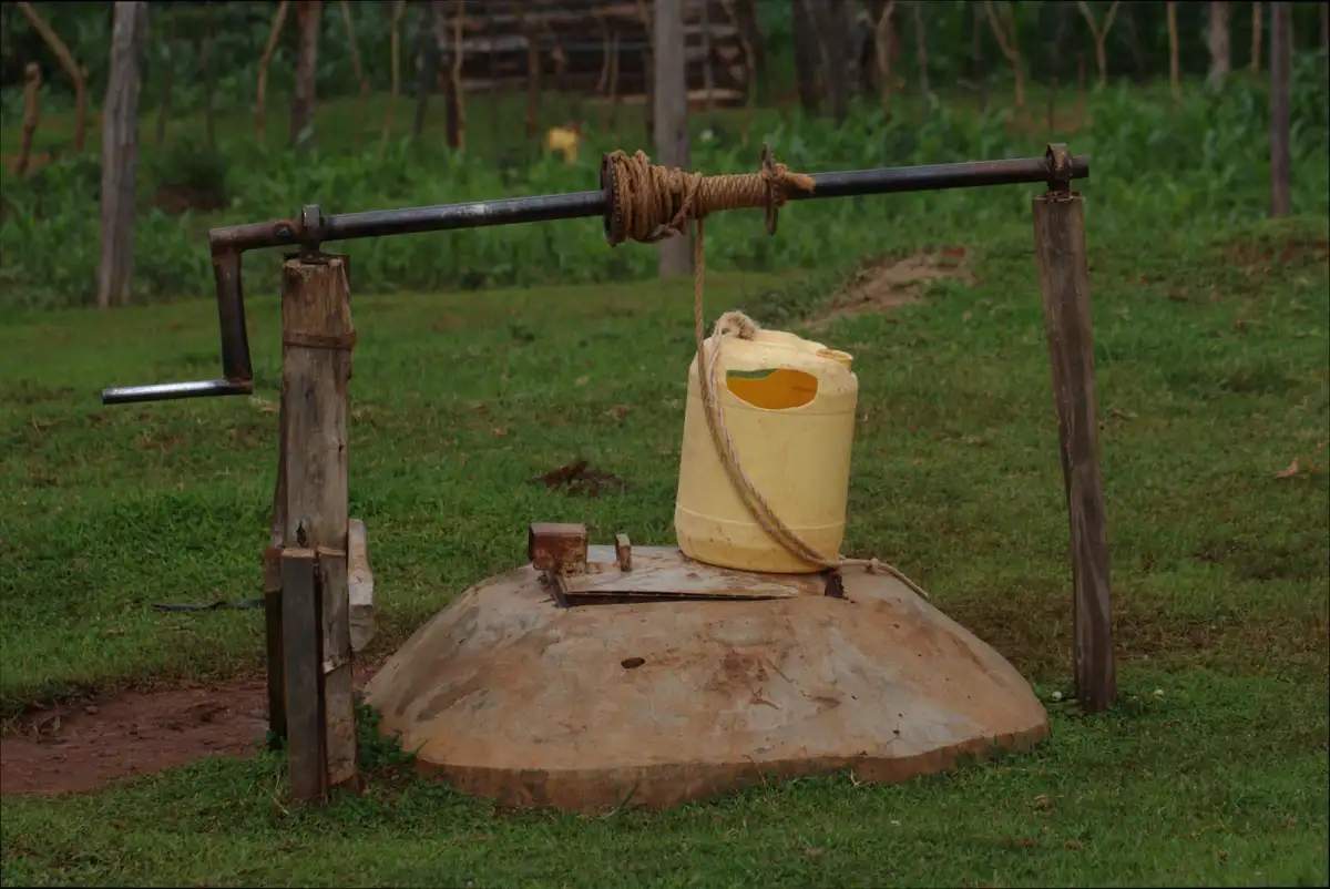 A wooden hand pump with a long iron handle, a rope and a yellow jerrycan on a concrete cover in a green field