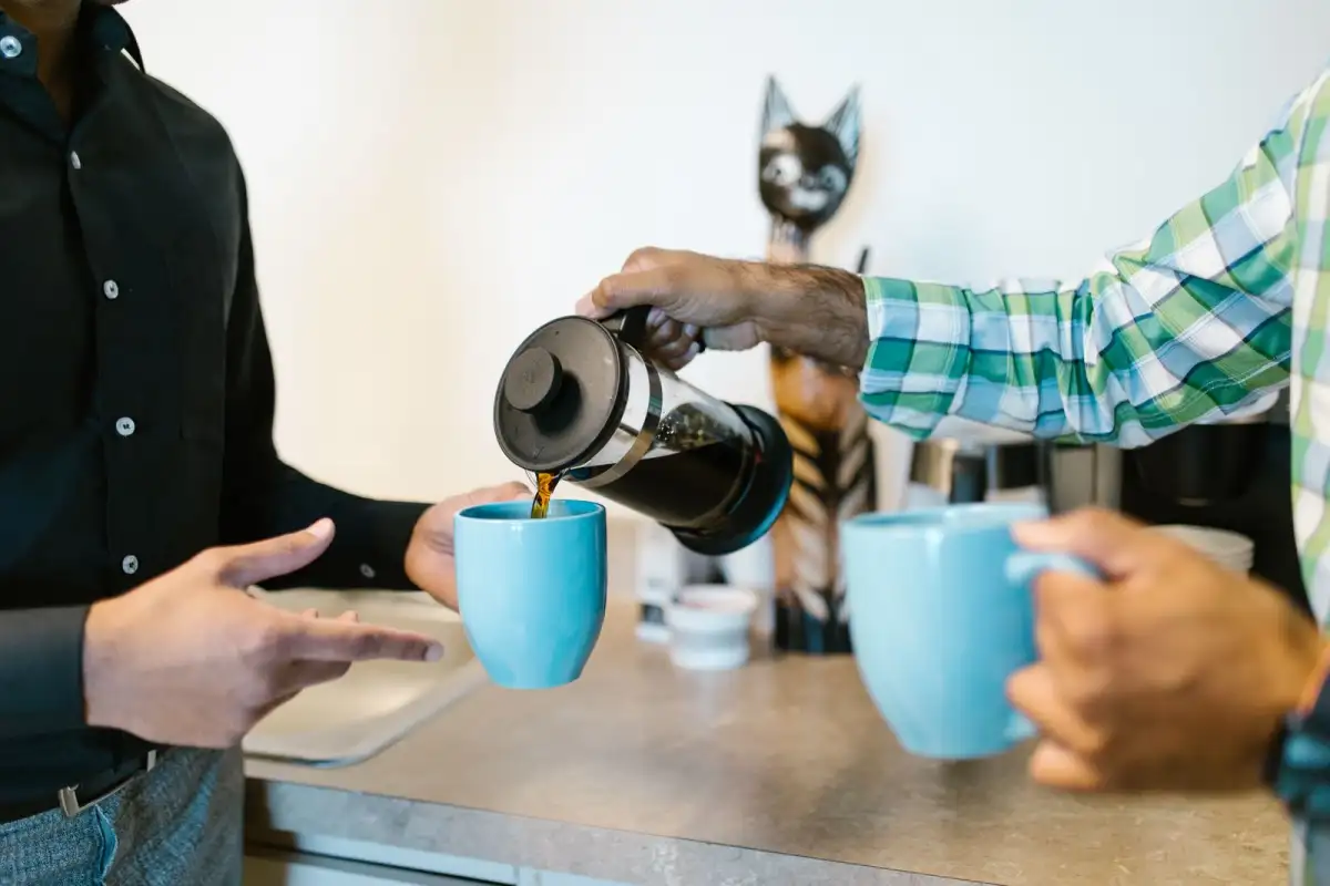 Two people pour coffee from a glass press into blue ceramic mugs at a counter