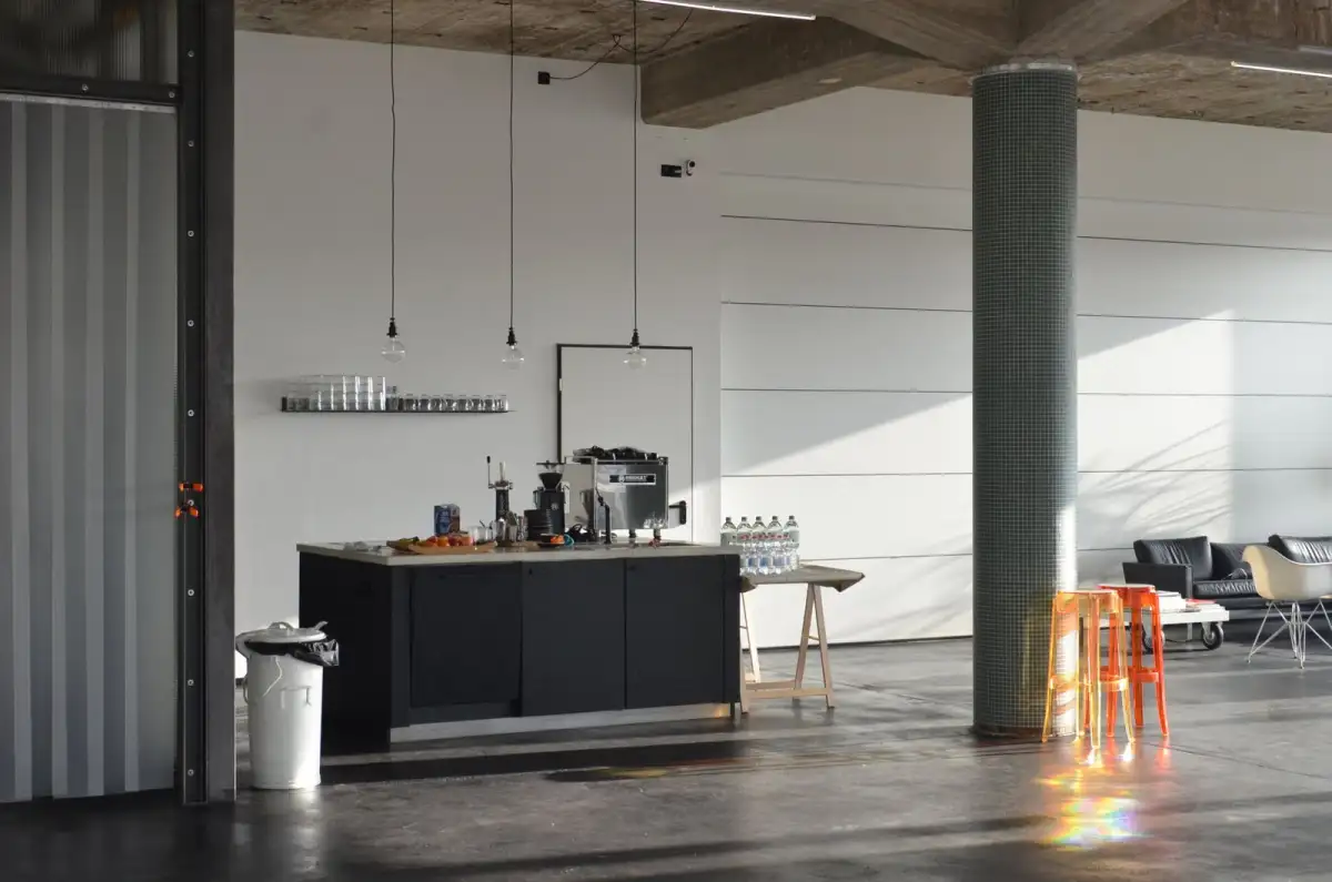 A concrete-floored kitchen area with a black counter, a coffee machine, water bottles, orange stools and a large column