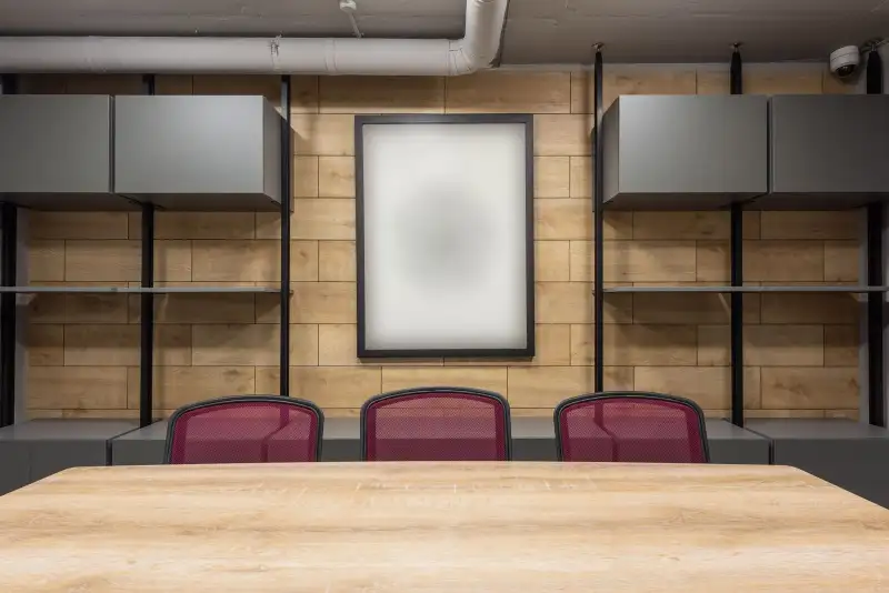 An empty boardroom with a pale wooden table, three burgundy chairs and a wood-panelled wall with a blank frame