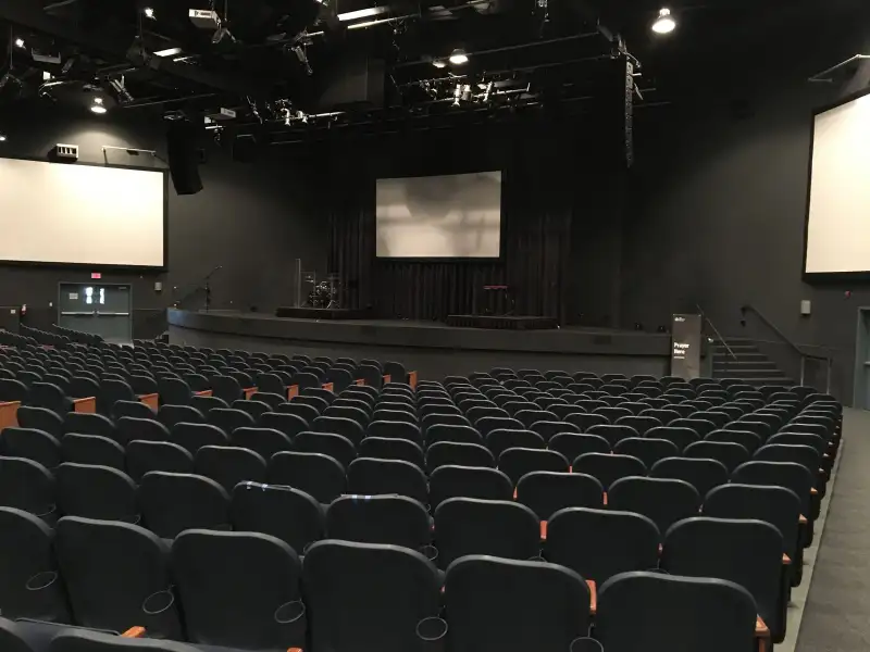 A dark auditorium with tiered rows of black seats facing a stage with screens and lighting rigs