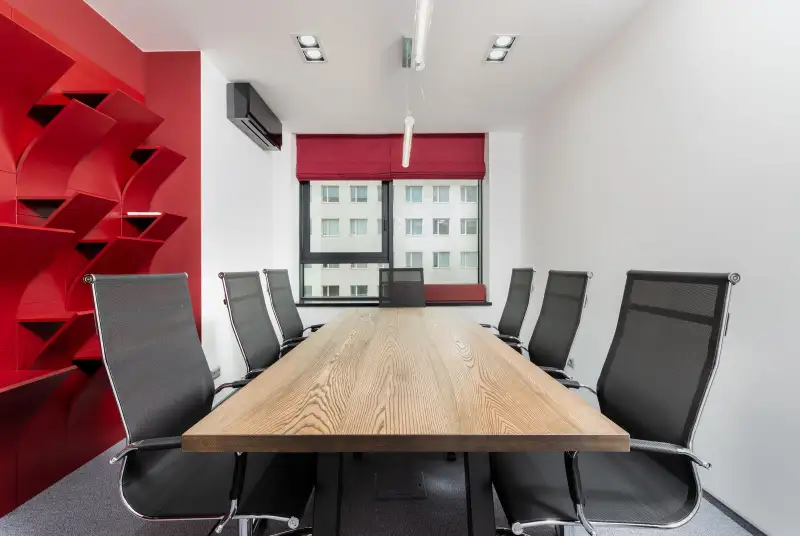 A meeting room with a long wooden table, black mesh chairs, a red shelving wall and a window