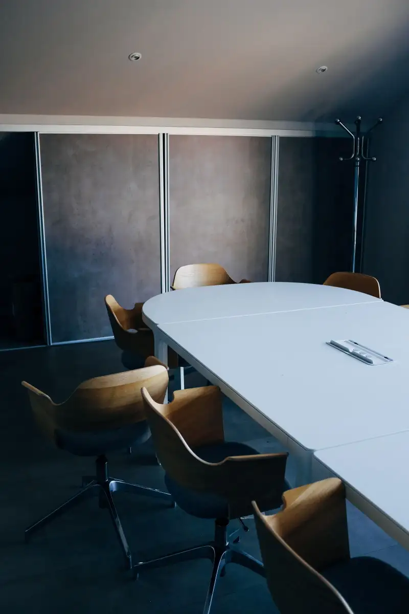 A small dark meeting room with a white oval table, wooden swivel chairs and a grey concrete-effect wall