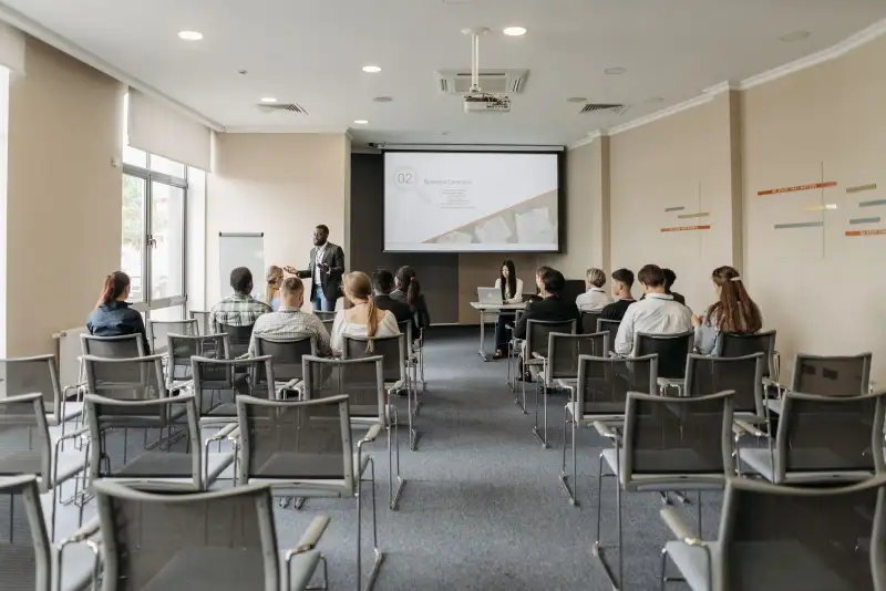 A workshop hall with rows of grey chairs facing a presenter and a projection screen, with people seated