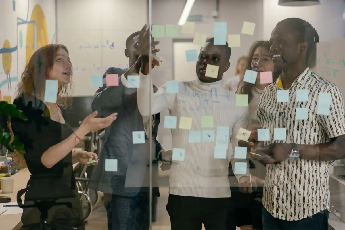 A group of colleagues stand at a glass wall covered in coloured sticky notes, laughing and pointing while they plan together