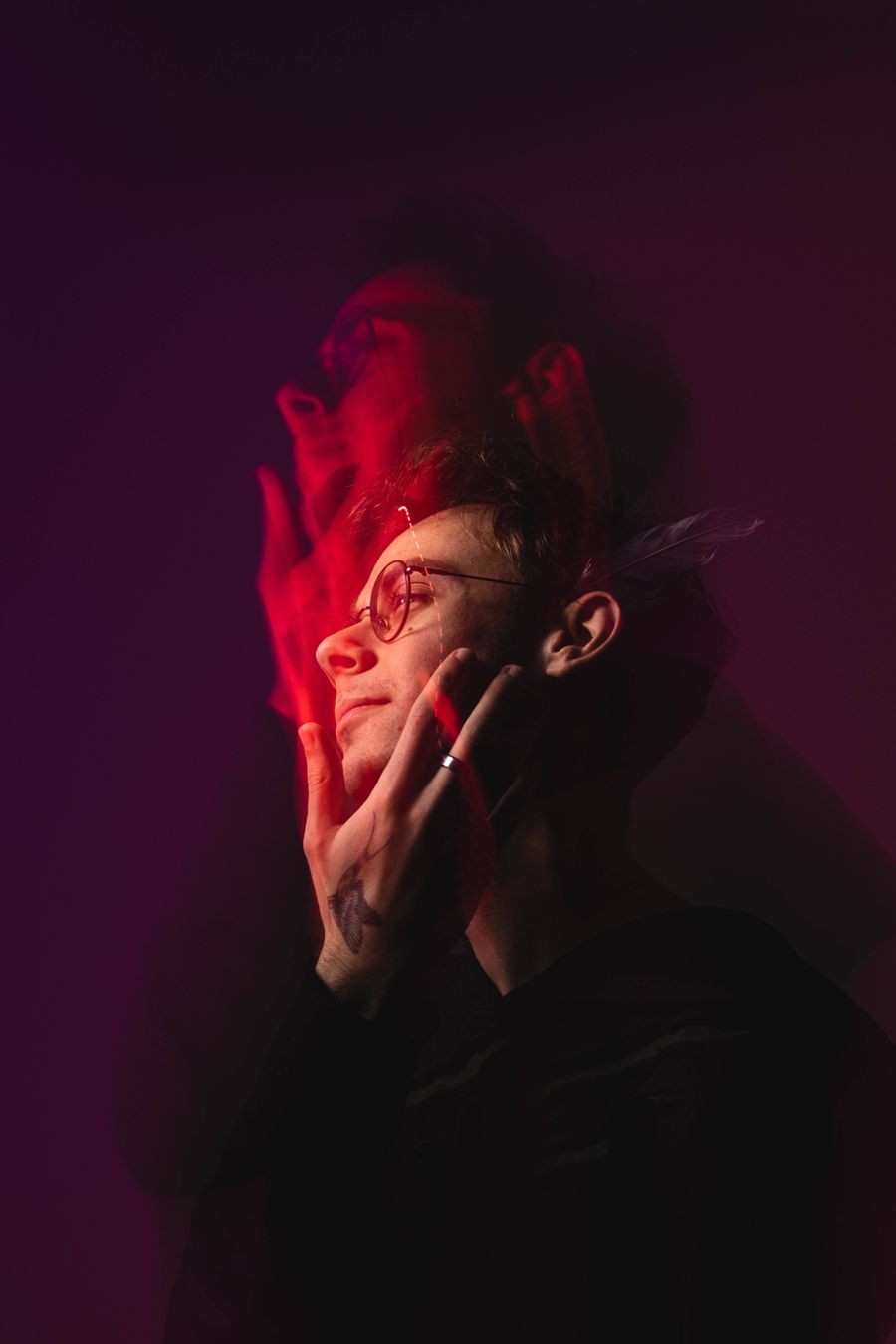 A young man in glasses with his hand on his face, lit in red and magenta