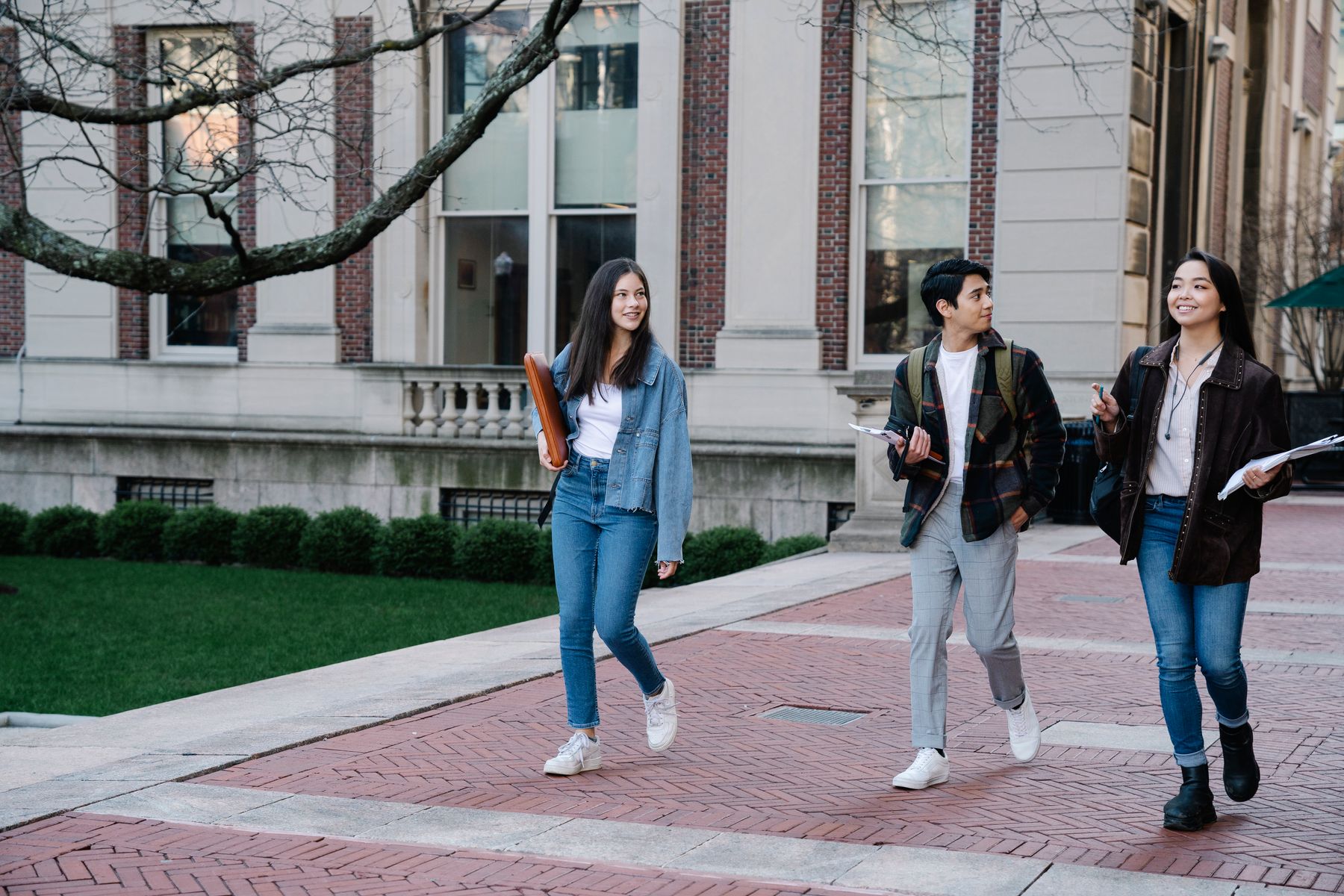 Three students in coats walking and talking on a brick path in front of a historic campus building
