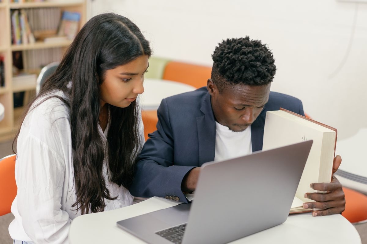Two students studying together over an open book and a laptop