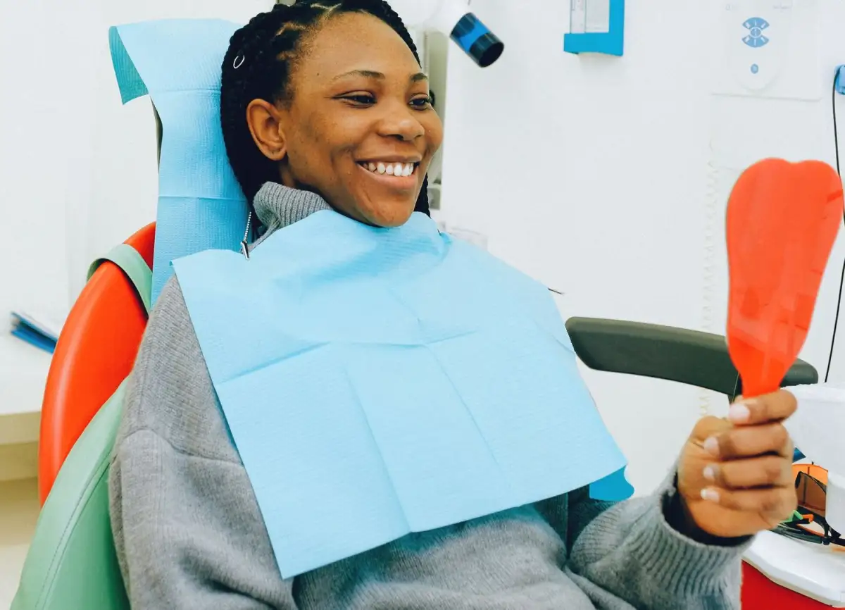 A smiling woman in a dental chair, with a paper bib, looks at herself in an orange hand mirror