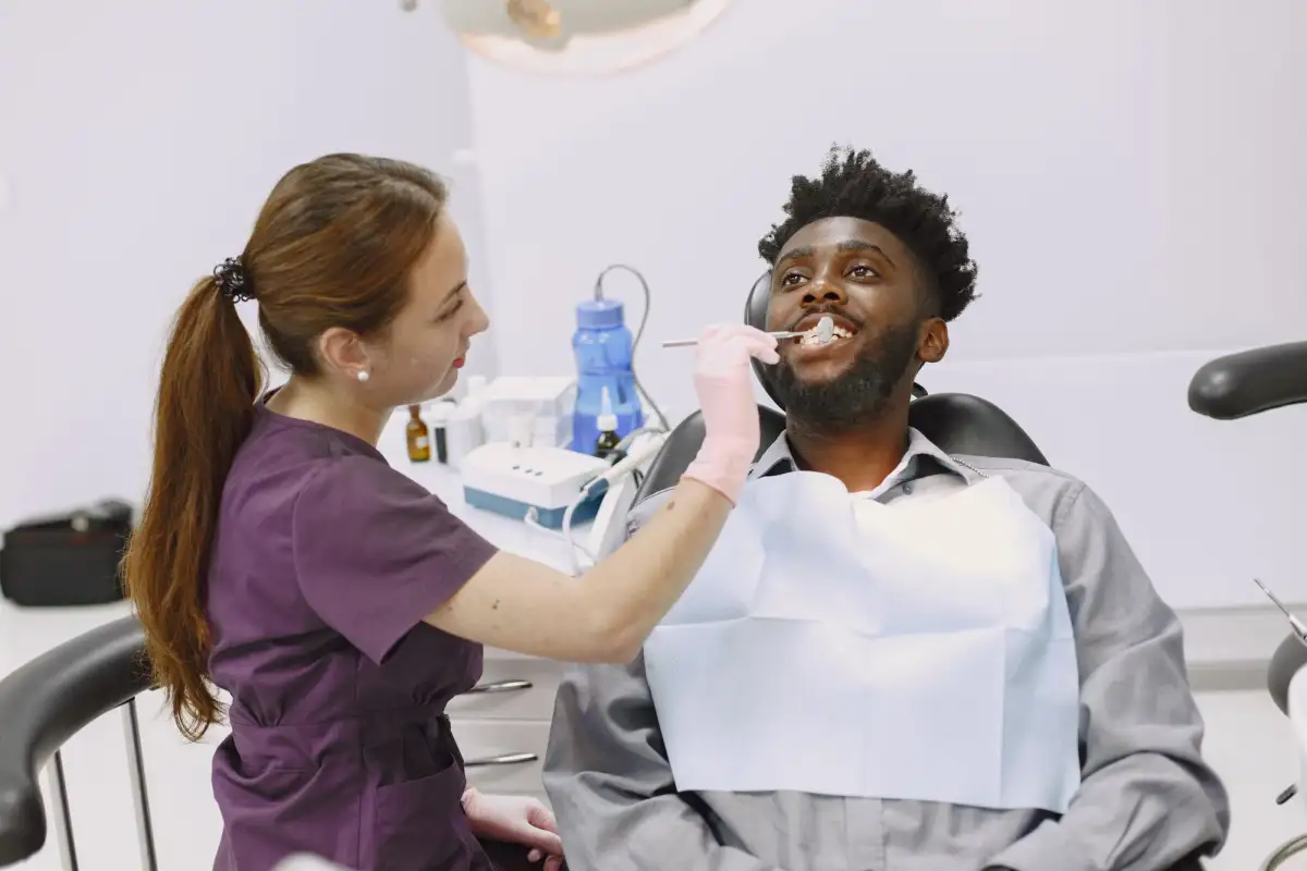 A dentist in purple scrubs and pink gloves examines a smiling patient's teeth with a small mirror