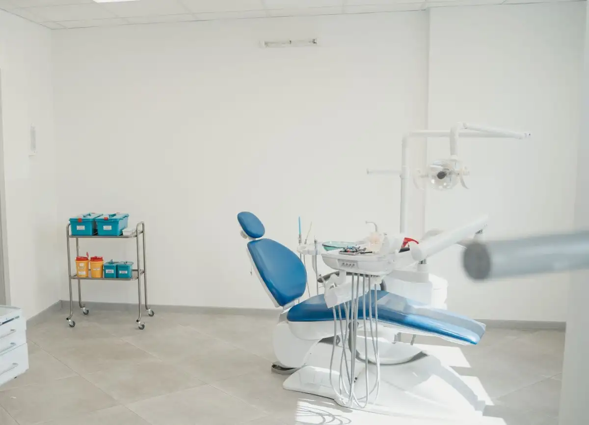 A bright treatment room with a blue dental chair, a sink and a trolley of sealed instrument boxes