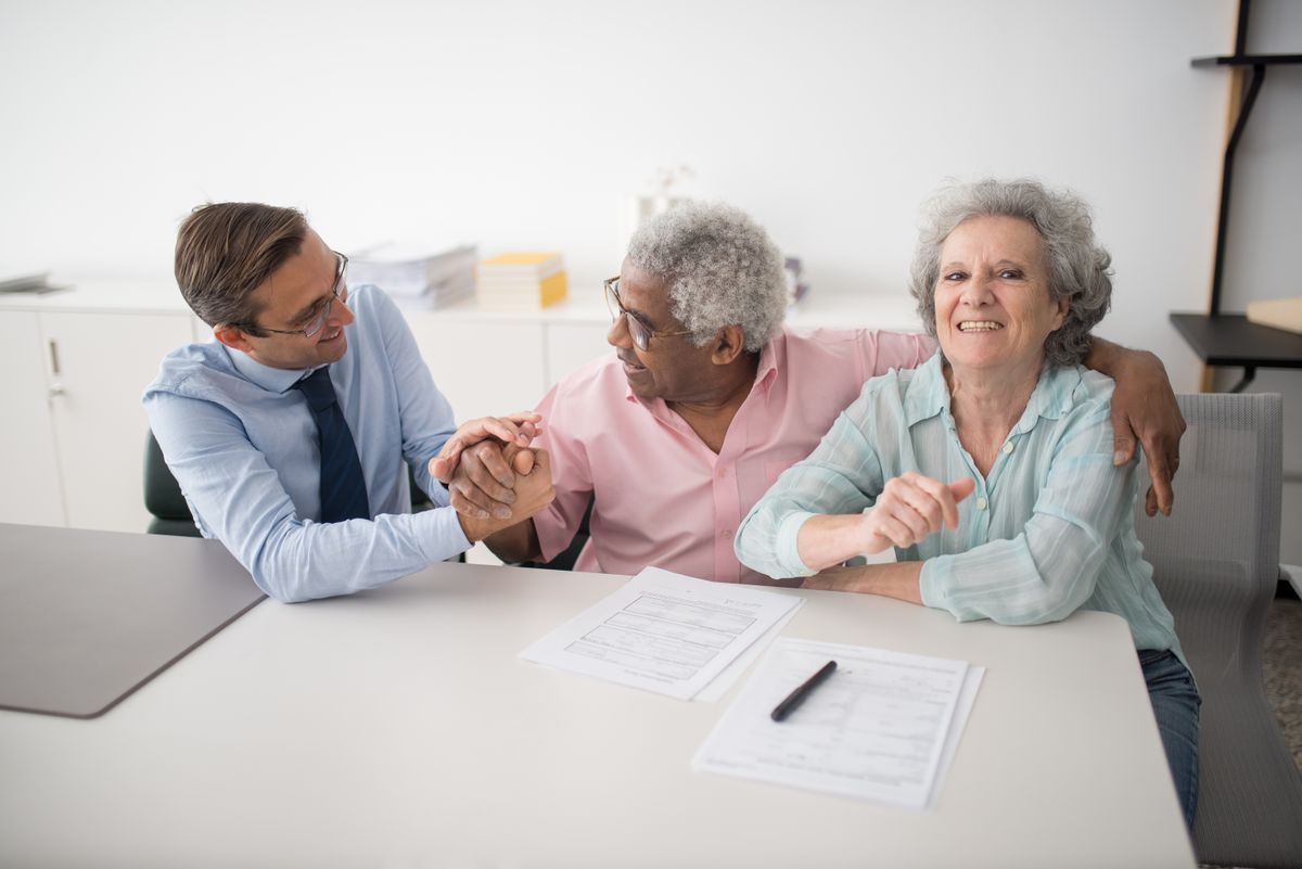 A smiling adviser holding the hand of an older client while another client looks on, with papers on the table