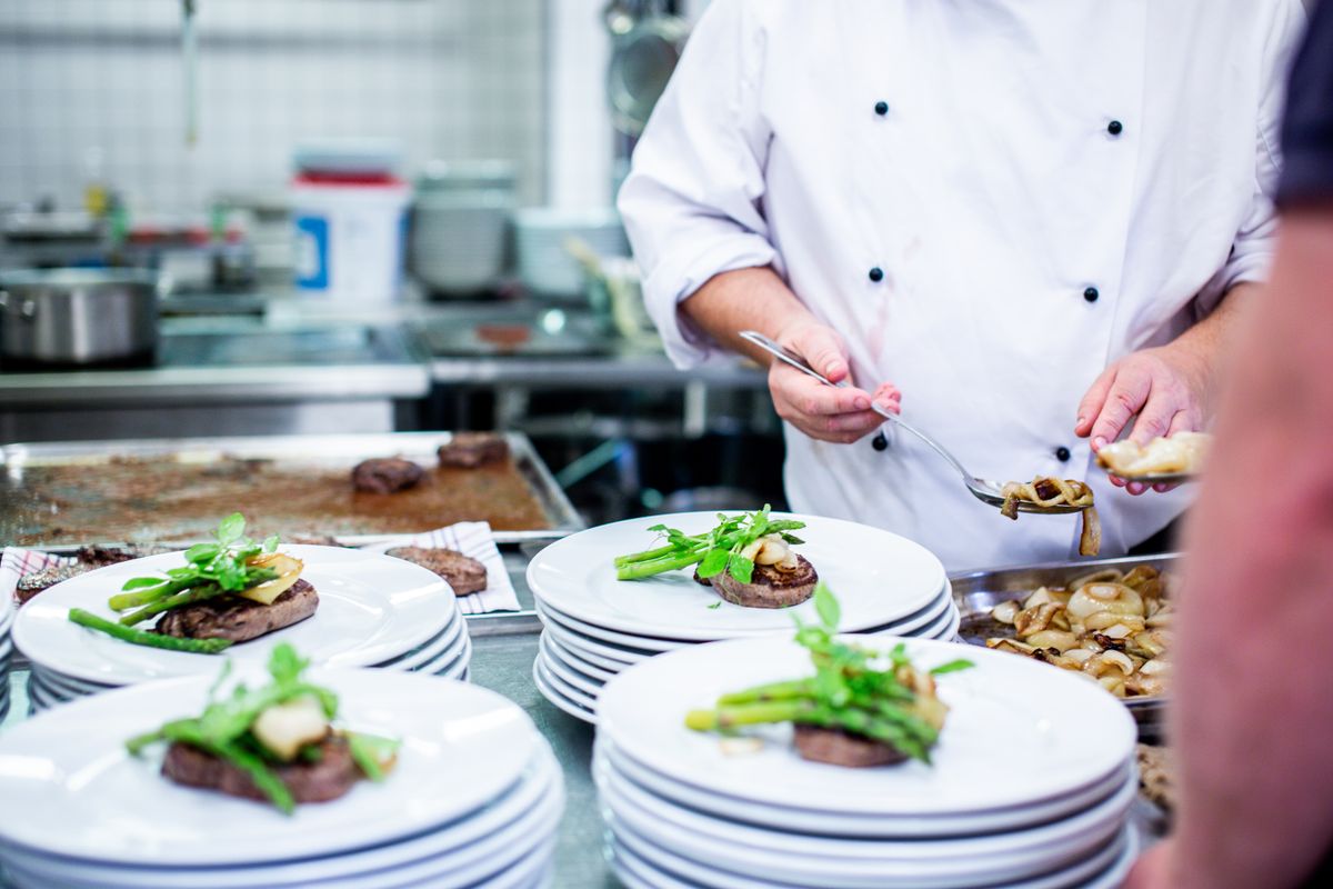 A chef in whites finishing plates of roast meat and greens on a kitchen pass