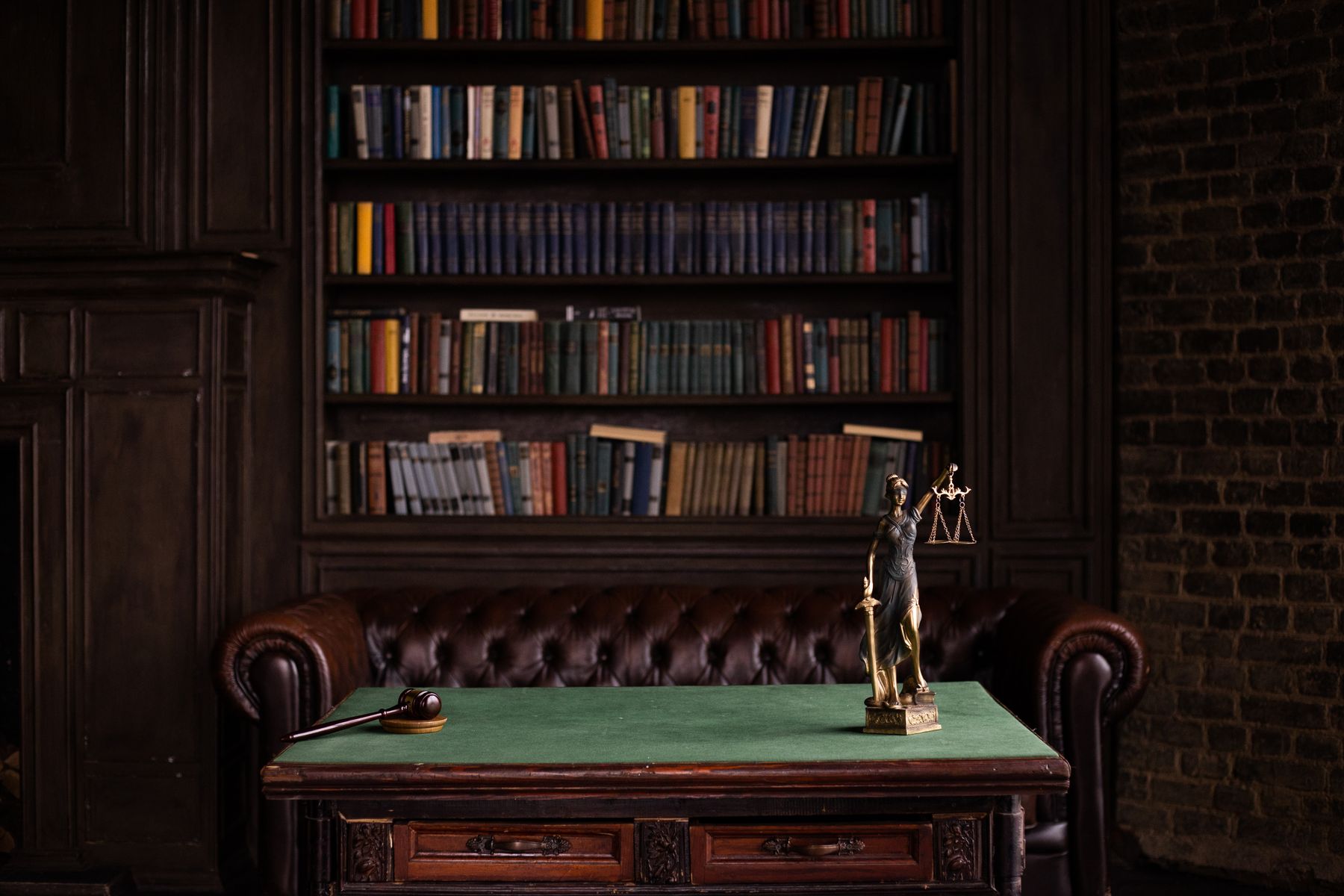 A dark wood-panelled law library with a leather chesterfield, a green-topped desk and a Lady Justice statue