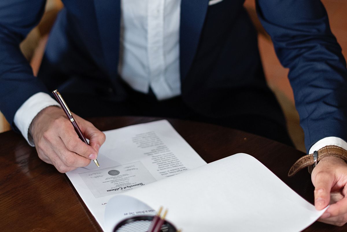A lawyer in a blue suit signing legal documents at a dark wooden table