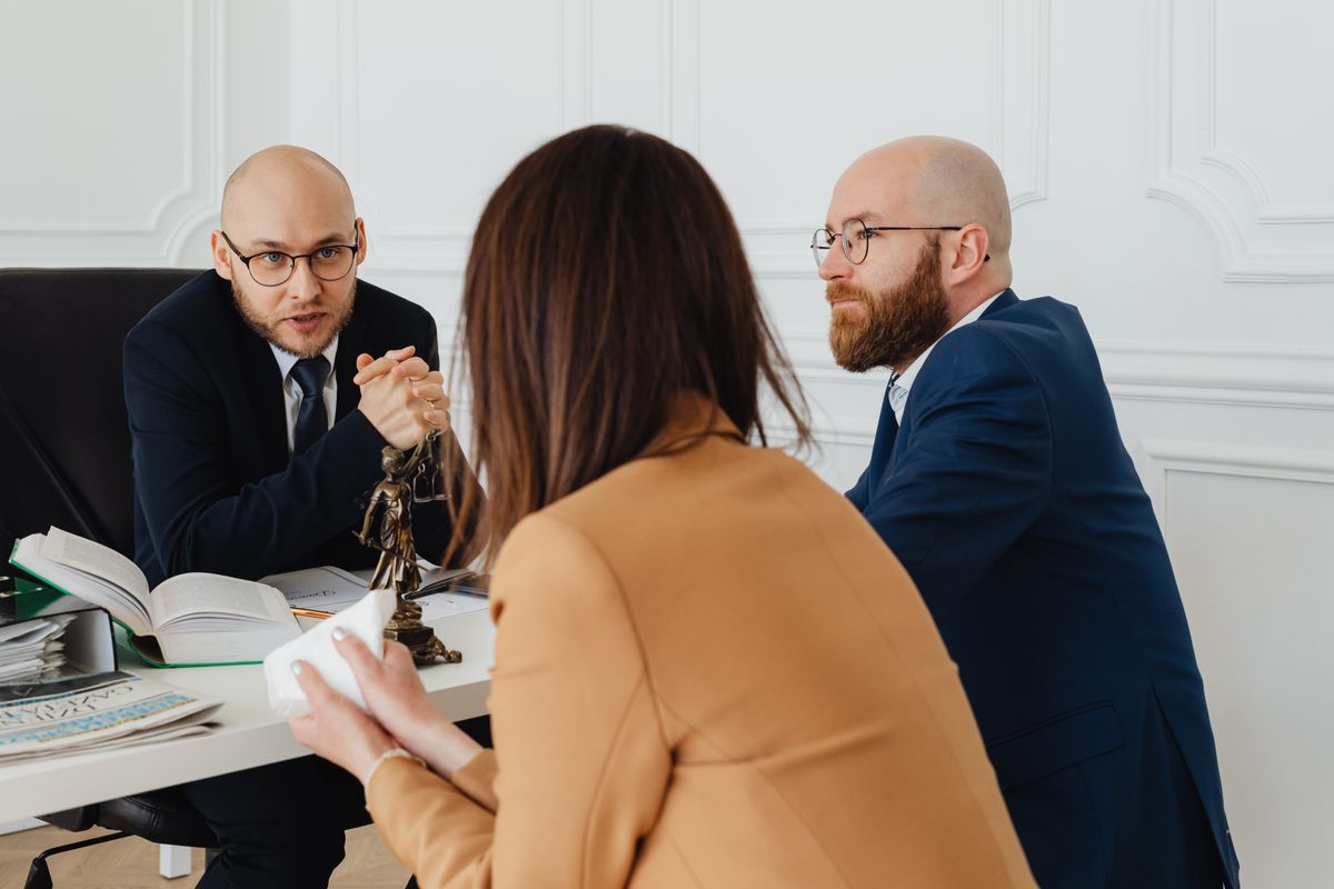 Two lawyers in dark suits advising a client at a table with a Lady Justice statuette