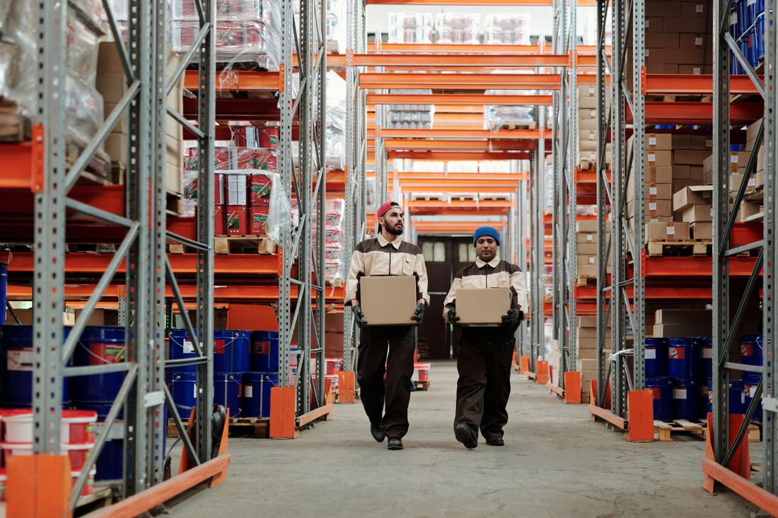 Two warehouse workers carrying parcels down a racked warehouse aisle