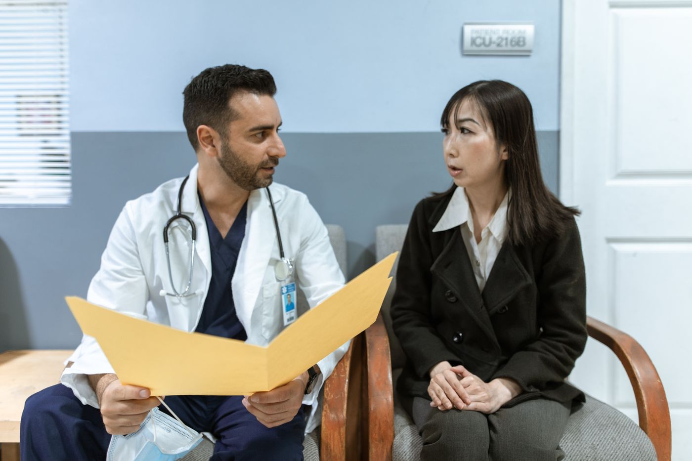 A doctor in a white coat holding a folder while speaking with a patient in a clinic waiting area