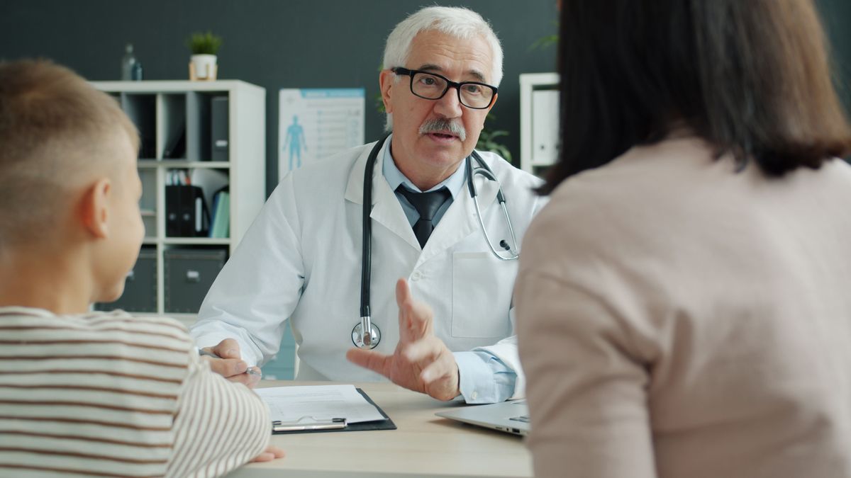 An older doctor in glasses speaking gently with a young boy and his mother
