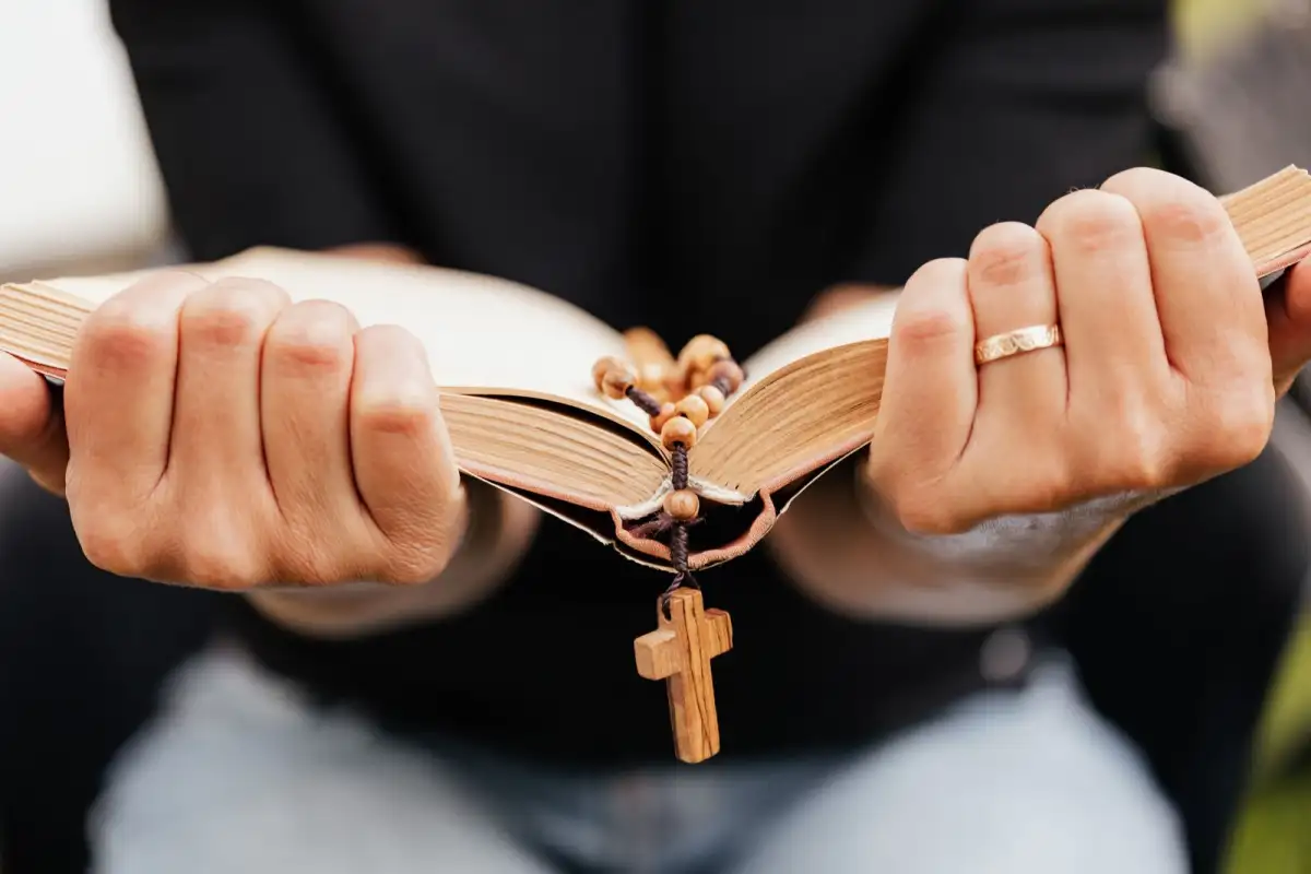 A person in dark clothes holds an old open bible in both hands, with a wooden cross on a rosary hanging beneath it