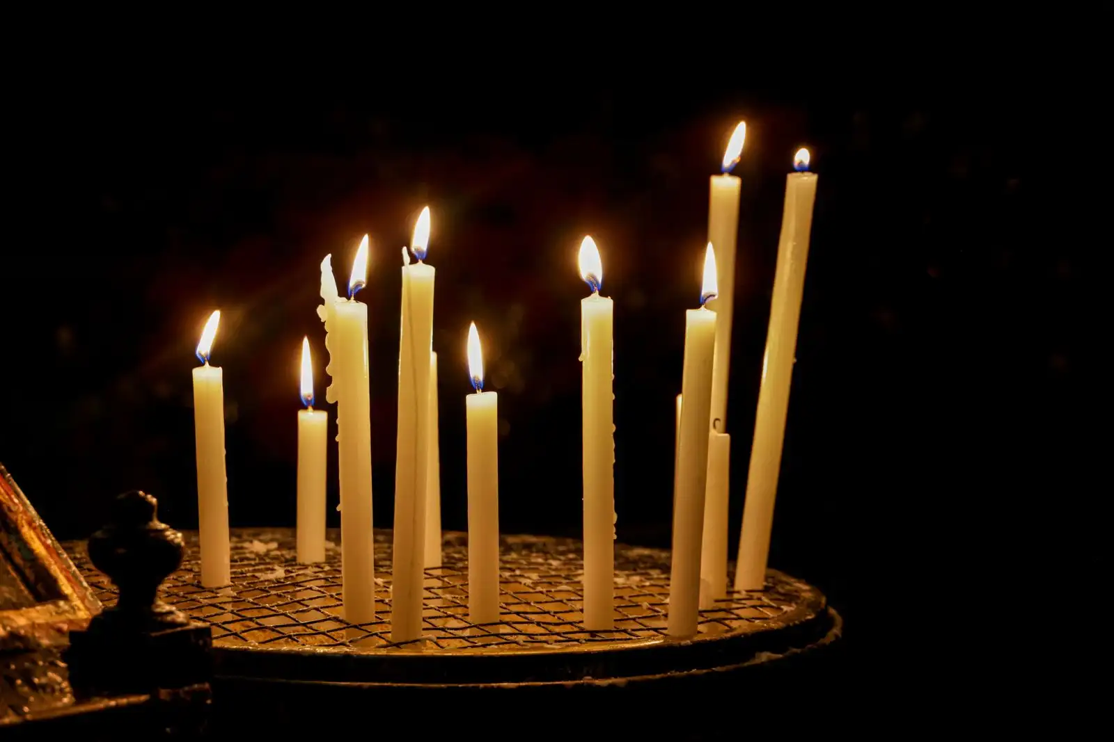 A dozen tall cream candles burning on a small round iron stand in the dark of a church