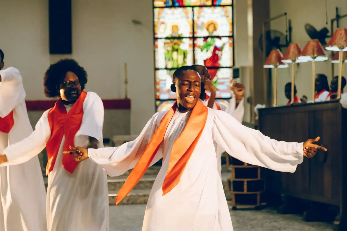 A choir in white robes with orange stoles sing joyfully in a church with a stained glass window behind them