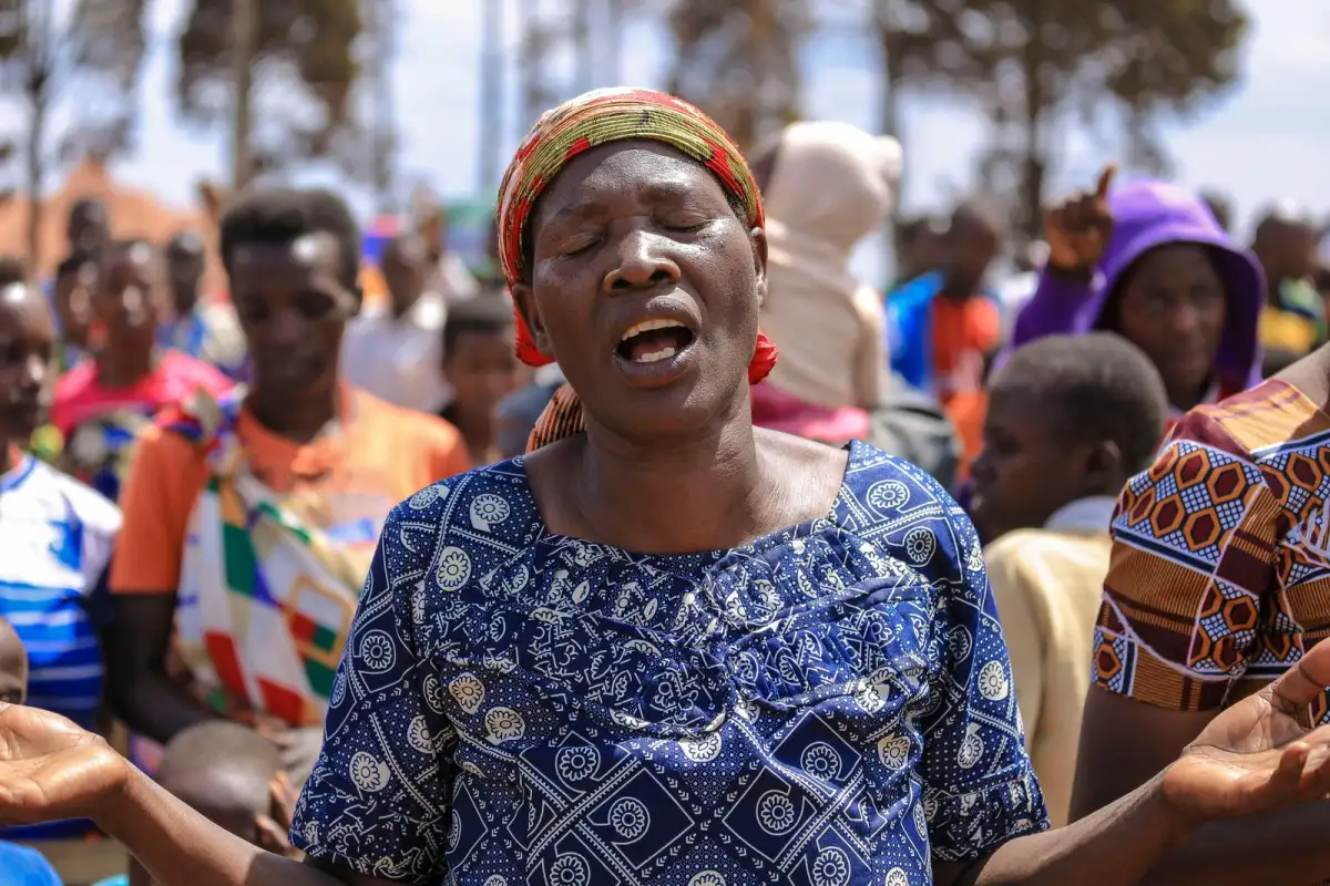 A woman with a bright headscarf and a blue patterned dress sings with her eyes closed and her arms open among a crowd of worshippers outdoors