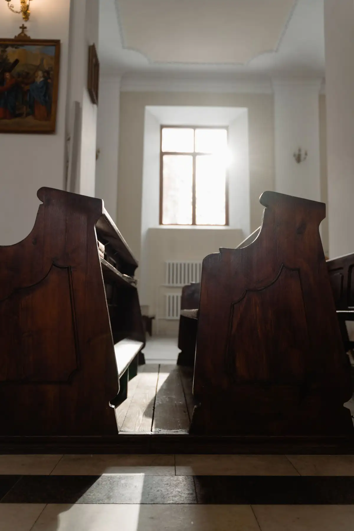 Two rows of dark wooden church pews face a plain window with sunlight streaming in, above a black and white tiled floor
