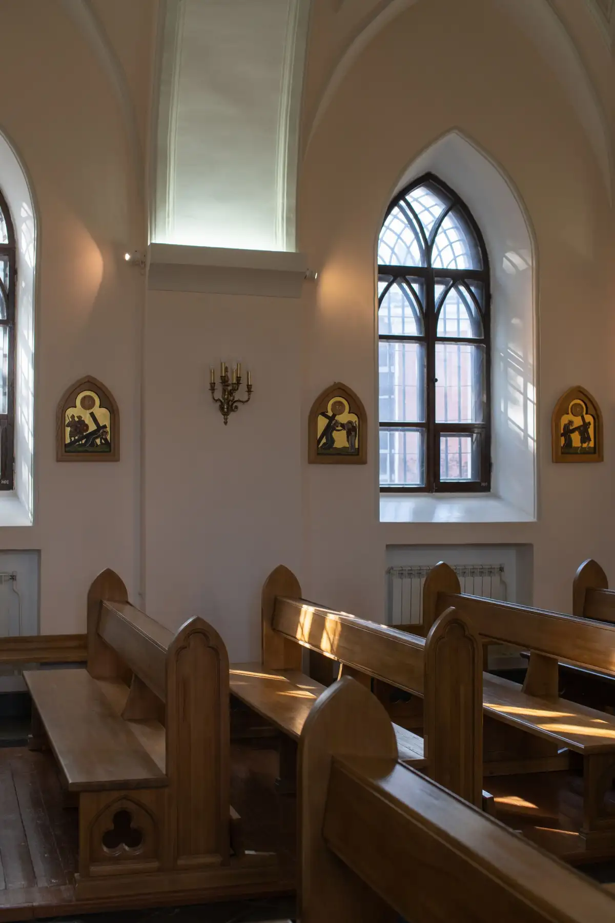 Warm wooden pews below a tall arched window, with framed pictures on the wall of a quiet church