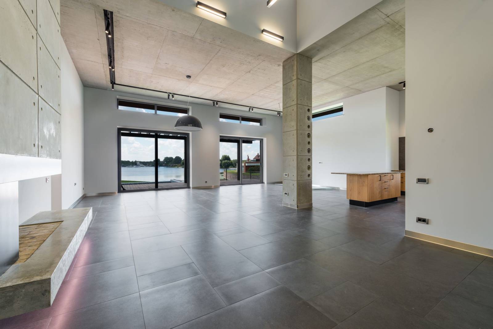 A tall concrete-ceilinged living space with a perforated concrete column and sliding glass doors opening to a lake